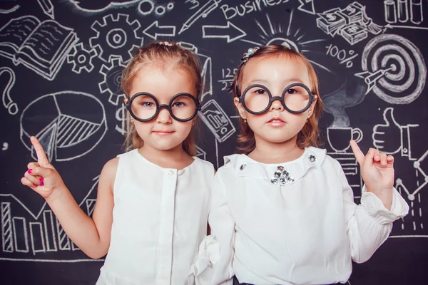 两个戴着眼镜的小女孩举起手指在墙上，墙上有商业或学校的照片 — 图库照片