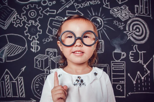 戴着眼镜的小聪明女孩用手指举在墙上的背景上，上面画着商业或学校的照片 — 图库照片