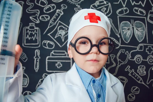 Niño feliz como médico en gafas con jeringa sobre fondo oscuro con patrón — Foto de Stock