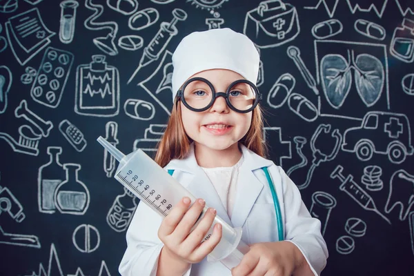 Happy little girl in doctor costume in glasses holding syringe on dark background with pattern — Stock Photo, Image