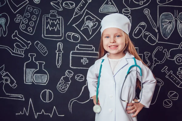 Niña con traje de médico sosteniendo el estetoscopio. Fondo oscuro con patrón. Sus manos en el cinturón — Foto de Stock