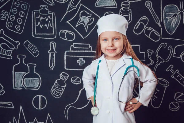 Niña con traje de médico sosteniendo el estetoscopio. Fondo oscuro con patrón. Sus manos en el cinturón — Foto de Stock