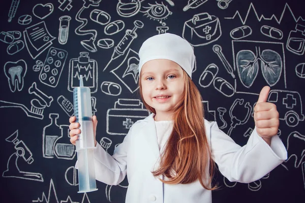 Niña con traje de médico sosteniendo la jeringa y muestra clase sobre fondo oscuro con patrón — Foto de Stock