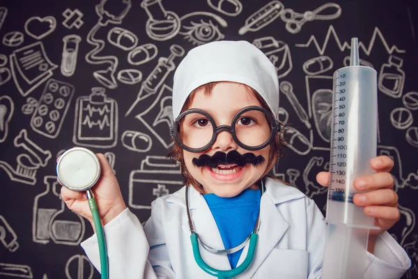 Niño feliz en traje médico sosteniendo jeringa y estetoscopio sobre fondo oscuro con patrón. El niño tiene bigote, gafas — Foto de Stock