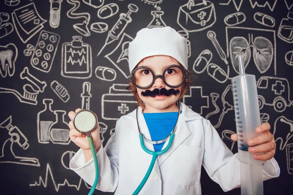 Niño feliz en traje médico sosteniendo jeringa y estetoscopio sobre fondo oscuro con patrón. El niño tiene bigote, gafas — Foto de Stock