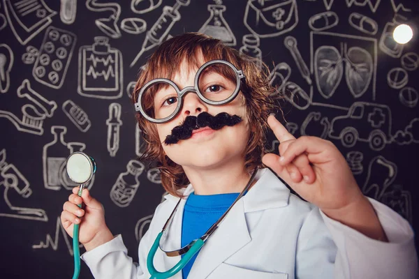 Niño feliz en traje de médico sosteniendo estetoscopio sobre fondo oscuro con patrón. Levanta pulgares hacia arriba. El niño tiene bigote, gafas — Foto de Stock