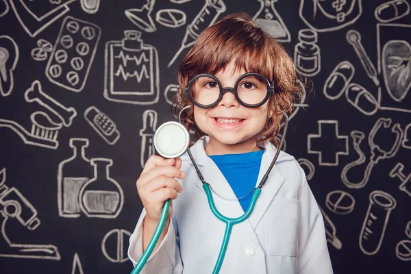 Niño feliz en traje de médico sosteniendo estetoscopio sobre fondo oscuro con patrón. El niño tiene gafas — Foto de Stock