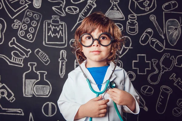 Niño feliz en traje de médico sosteniendo estetoscopio sobre fondo oscuro con patrón. El niño tiene gafas — Foto de Stock