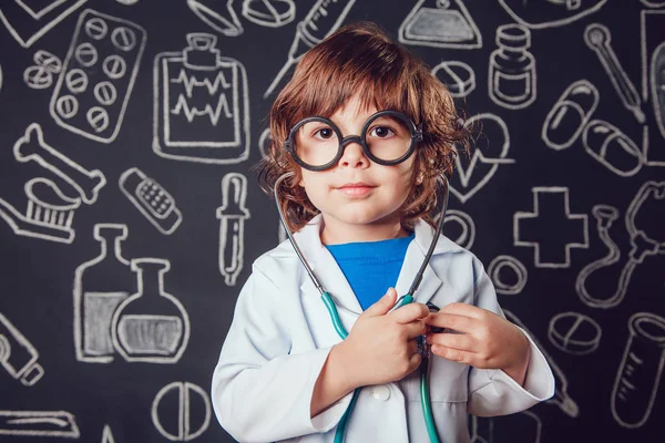 Niño feliz en traje de médico sosteniendo estetoscopio sobre fondo oscuro con patrón. El niño tiene gafas — Foto de Stock