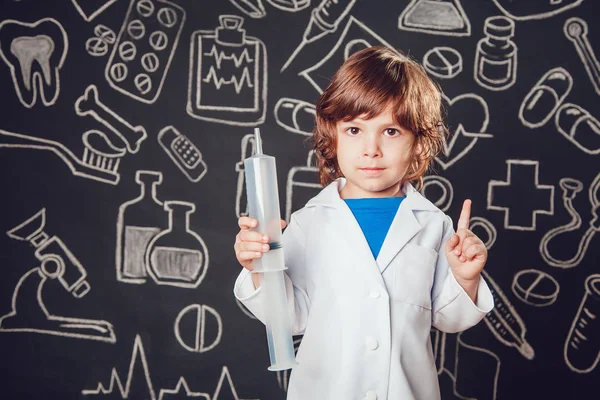 Niño feliz disfrazado de doctor sosteniendo la jeringa sobre fondo oscuro con patrón. Levanta pulgares hacia arriba . — Foto de Stock