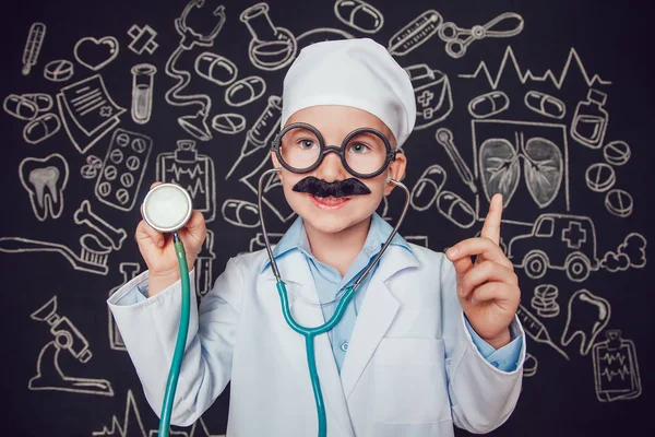 Niño en traje de médico sosteniendo estetoscopio sobre fondo oscuro con patrón. Levanta pulgares hacia arriba. El niño tiene bigote y gafas — Foto de Stock