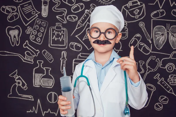Little boy in doctor costume holding syringe on dark background with pattern. Lifts thumbs up. The child has mustache and glasses — Stock Photo, Image
