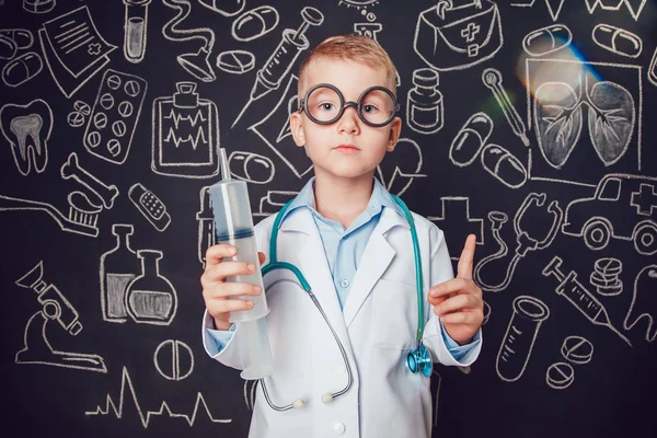 Little boy in doctor costume holding syringe on dark background with pattern. Lifts thumbs up. The child has glasses — Stock Photo, Image