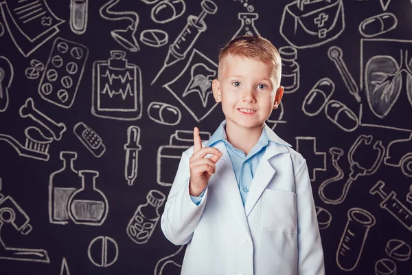 Happy little boy in doctor costume standing and on dark background with pattern. The child has glasses — Stock Photo, Image