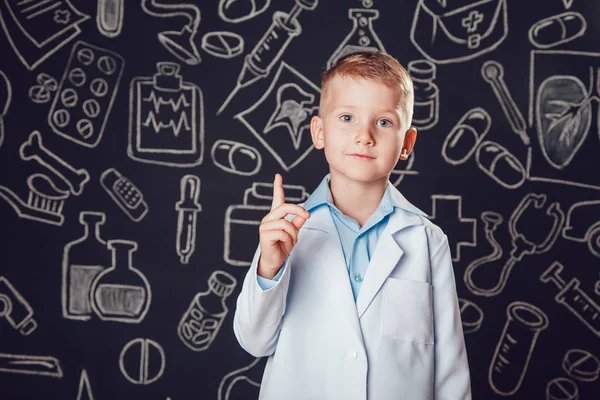 Niño en traje de médico de pie y levanta pulgares hacia arriba sobre fondo oscuro con patrón . — Foto de Stock