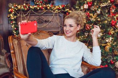 Genç sarışın kadın Noel ağacı yakın bir sandalyede oturan güzel ve selfie yapmak.