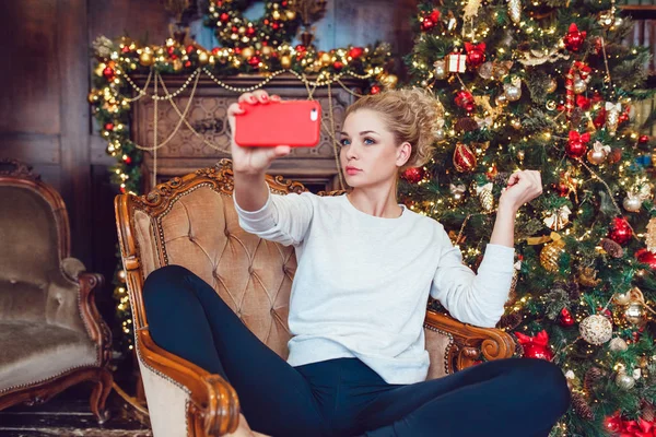 Junge hübsche blonde Frau sitzt auf einem Stuhl in der Nähe des Weihnachtsbaums und macht Selfie. — Stockfoto