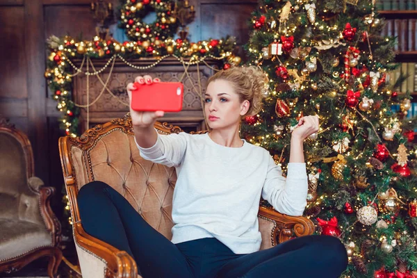 Junge hübsche blonde Frau sitzt auf einem Stuhl in der Nähe des Weihnachtsbaums und macht Selfie. — Stockfoto