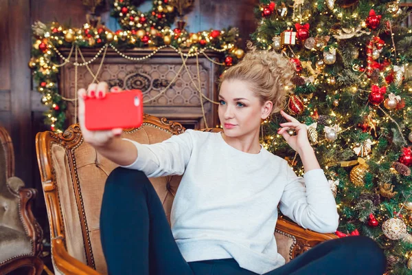 Junge hübsche blonde Frau sitzt auf einem Stuhl in der Nähe des Weihnachtsbaums und macht Selfie. — Stockfoto