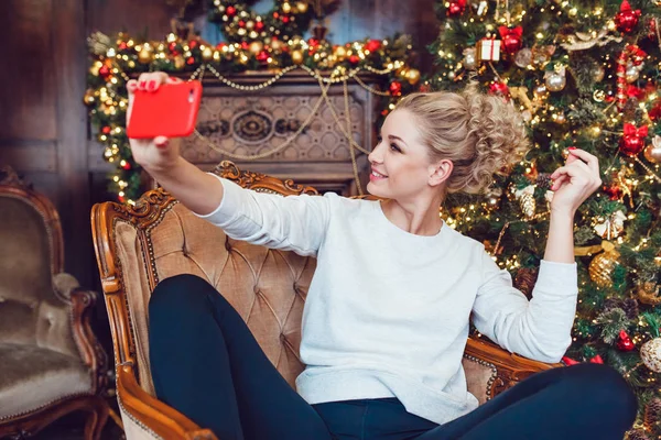 Junge hübsche blonde Frau sitzt auf einem Stuhl in der Nähe des Weihnachtsbaums und macht Selfie. — Stockfoto