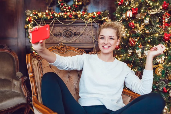 Junge hübsche blonde Frau sitzt auf einem Stuhl in der Nähe des Weihnachtsbaums und macht Selfie. — Stockfoto