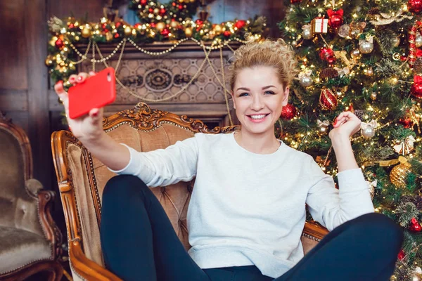 Junge hübsche blonde Frau sitzt auf einem Stuhl in der Nähe des Weihnachtsbaums und macht Selfie. — Stockfoto