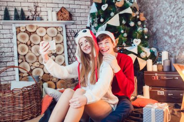 Genç çift oturma komik şapkalar eğleniyor Noel ağacı yakınındaki aşk ve selfie yapmak. Yeni yıl