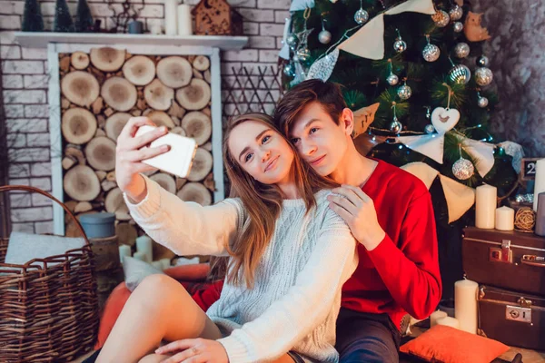 Jovem casal apaixonado sentado perto da árvore de natal se divertindo e fazer selfie. Ano Novo — Fotografia de Stock