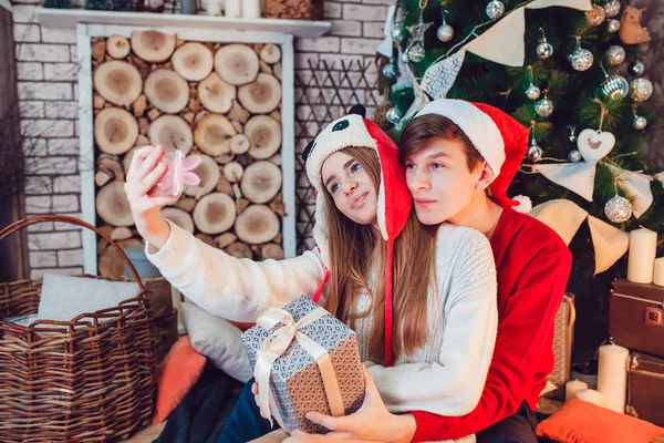 Young couple in love sitting near christmas tree in funny hats having fun and make selfie. New Year — Stock Photo, Image