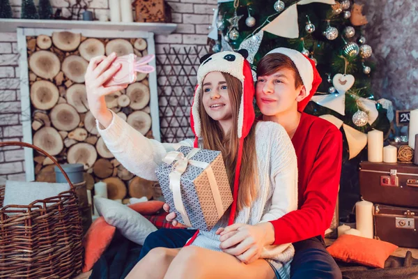 Young couple in love sitting near christmas tree in funny hats having fun and make selfie. New Year — Stock Photo, Image