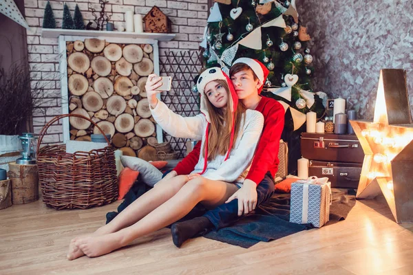 Young couple in love sitting near christmas tree in funny hats having fun and make selfie. New Year — Stock Photo, Image