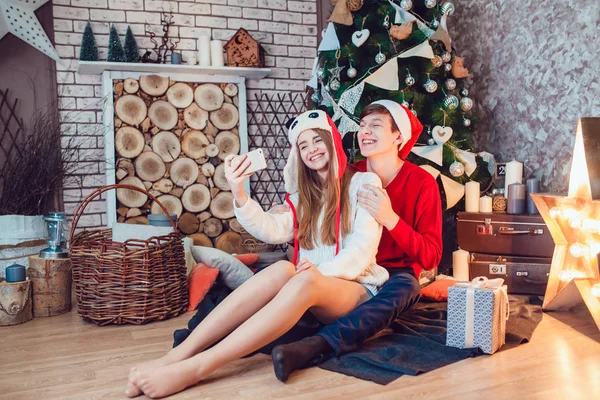 Jovem casal apaixonado sentado perto da árvore de natal em chapéus engraçados se divertindo e fazer selfie. Ano Novo — Fotografia de Stock