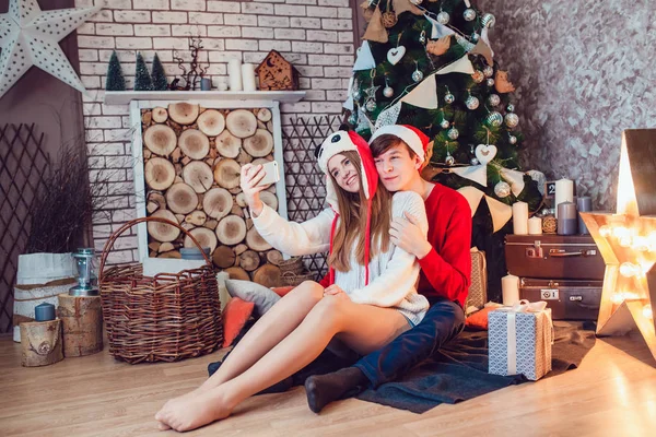 Jovem casal apaixonado sentado perto da árvore de natal em chapéus engraçados se divertindo e fazer selfie. Ano Novo — Fotografia de Stock