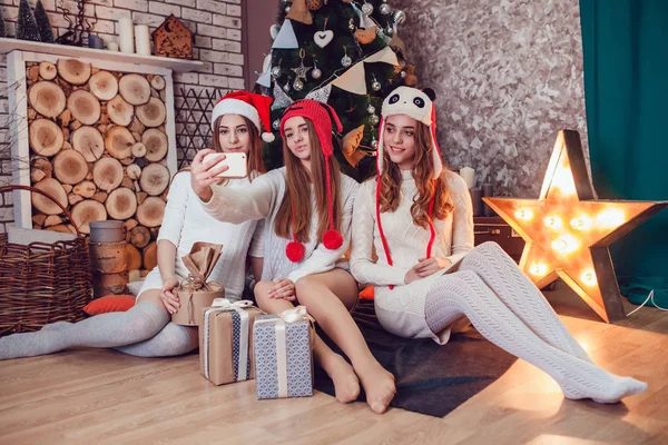 Meninas bonitas em chapéus engraçados sentados no chão perto de uma árvore de Natal com presentes e fazer selfie. Ano Novo . — Fotografia de Stock