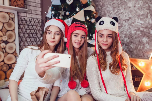 Meninas bonitas em chapéus engraçados sentados no chão perto de uma árvore de Natal e fazer selfie. Ano Novo . — Fotografia de Stock