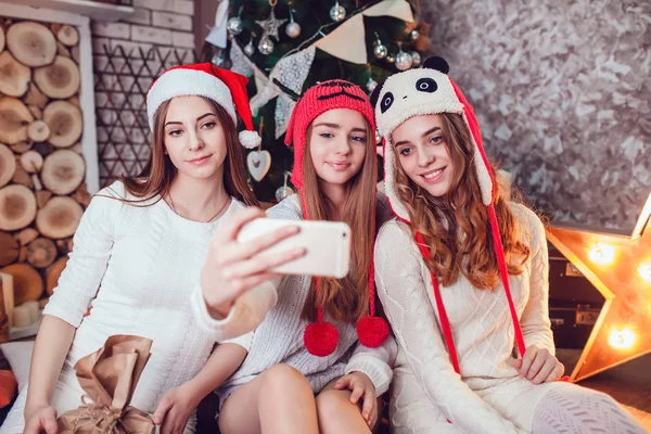 Meninas bonitas em chapéus engraçados sentados no chão perto de uma árvore de Natal e fazer selfie. Ano Novo . — Fotografia de Stock