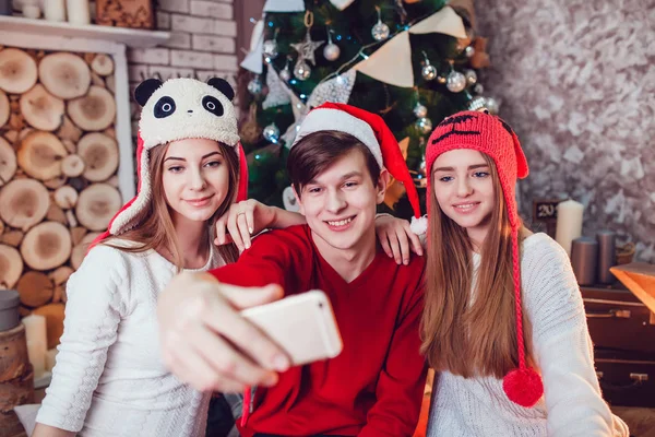 Meninas bonitas e menino em chapéus engraçados sentado no chão perto de uma árvore de Natal com presentes. Faz selfie. Ano Novo . — Fotografia de Stock