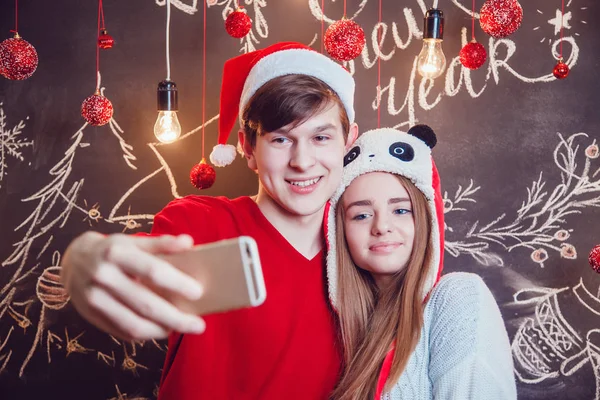 Casal feliz em chapéus engraçados de pé abraçando e fazer selfie em um fundo escuro com padrão de Natal. Ano Novo . — Fotografia de Stock