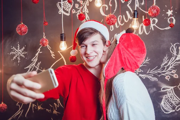 Casal feliz em chapéus engraçados de pé abraçando e fazer selfie em um fundo escuro com padrão de Natal. Ano Novo. A menina beija cara . — Fotografia de Stock