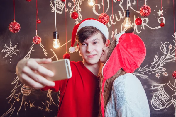 Casal feliz em chapéus engraçados de pé abraçando e fazer selfie em um fundo escuro com padrão de Natal. Ano Novo. A menina beija cara . — Fotografia de Stock