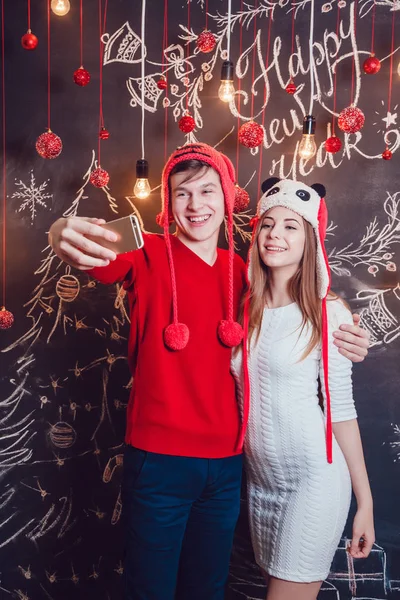 Casal feliz em chapéus engraçados de pé abraçando e fazer selfie em um fundo escuro com padrão de Natal. Ano Novo . — Fotografia de Stock