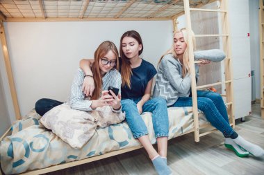 Beautiful girls sitting on a bed in hostel talk and using smartphone. clipart