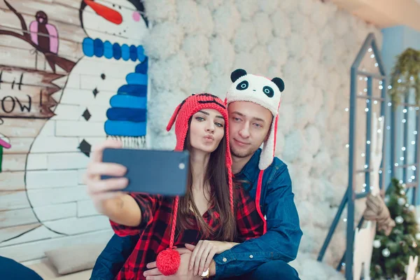 Hermosa pareja joven en sombreros divertidos sentado en el fondo de las decoraciones de Navidad y hacer selfie . —  Fotos de Stock