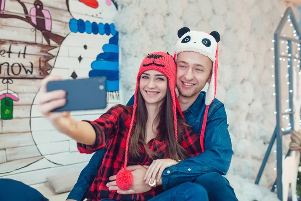 Hermosa pareja joven en sombreros divertidos sentado en el fondo de las decoraciones de Navidad y hacer selfie . —  Fotos de Stock