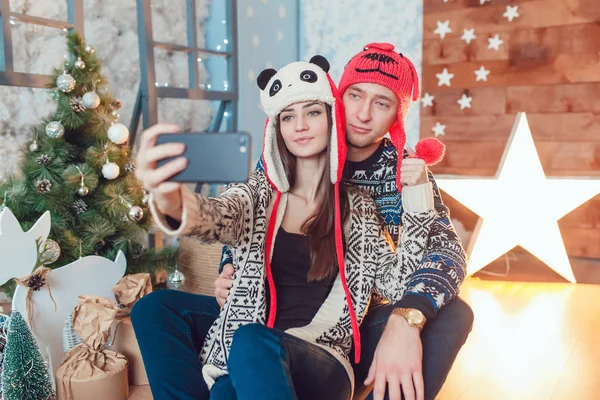 Bonito jovem casal em chapéus engraçados sentado no fundo de decorações de Natal e fazer selfie . — Fotografia de Stock