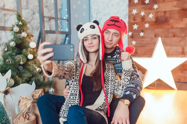 Beautiful young couple in funny hats sitting on the background of Christmas decorations and make selfie. — Stock Photo, Image