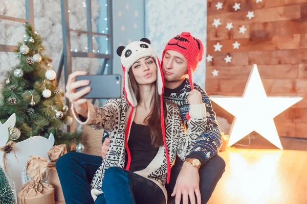 Bonito jovem casal em chapéus engraçados sentado no fundo de decorações de Natal e fazer selfie . — Fotografia de Stock