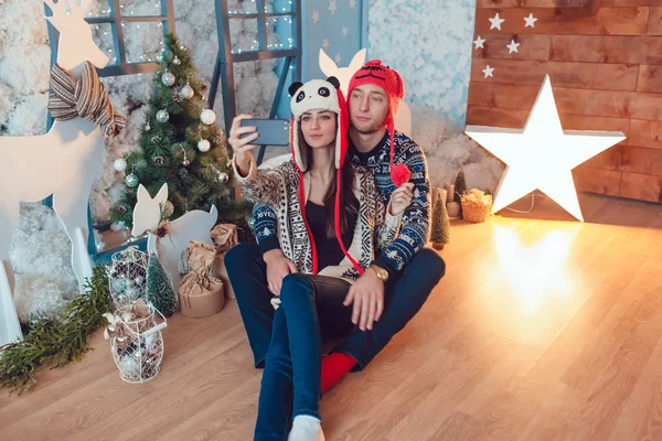 Vackra unga par i roliga hattar sitter på bakgrunden av juldekorationer och göra selfie. — Stockfoto
