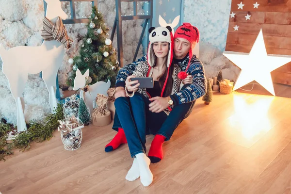 Beautiful young couple in funny hats sitting on the background of Christmas decorations and make selfie. — Stock Photo, Image