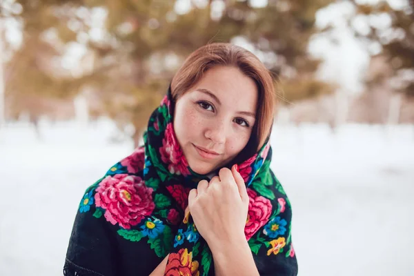 Russian beauty woman with red hair and freckles in the national scarf stands on a background of trees. — Stock Photo, Image